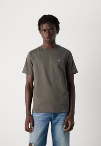Ung mand med korte dreadlocks iført en mørkegrå Calvin Klein t-shirt og lyseblå slidte jeans, stående mod en helt hvid baggrund.