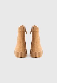 Une paire de bottines en daim beige avec des semelles en caoutchouc côtelées et des fermetures à glissière à l'arrière, présentée de l'arrière sur un fond blanc uni.