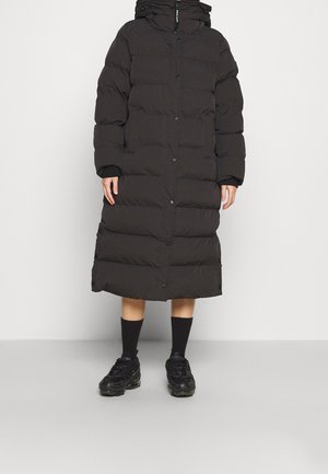 Personne portant un long manteau matelassé noir avec des boutons-pression, associé à des chaussettes noires et des baskets épaisses noires, se tenant sur un fond uni.