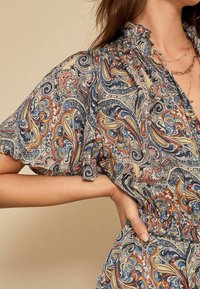 Femme portant un blouse à motifs paisley multicolores et une jupe assortie, la main reposant sur la hanche, portant des colliers en or superposés.