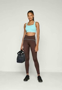 Lichtblauw sportcrop top, bruine hoge taille leggings met zijpiping, en zwarte sneakers; houdt een zwarte sporttas vast.