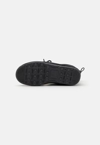 Svart sneakersåle med teksturert gummi, utstyrt med sirkulære mønstre for godt grep og en drakksløyfe bak for enkel påslipping.