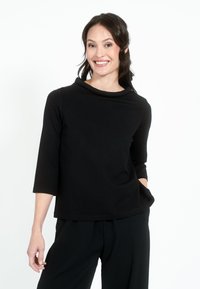 Črn top s tremi četrtinami rokavov, z vratnim izrezom v obliki kapuce in sproščenim krojem. Mehak material, minimalističen dizajn in vidne stranske žepe.