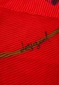 Tessuto rosso con una texture a coste; presenta una striscia verde accentuata e una firma nera "Lesquesual" tessuta nel tessuto.