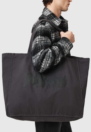 Osoba ubrana w czarno-białą kurtkę w kratę, nosząca dużą czarną torbę na ramię na jednolitym tle.