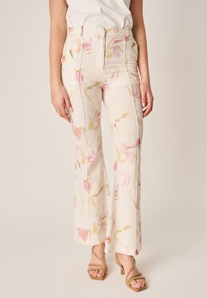 Donna che indossa pantaloni a vita alta e gamba larga color crema con stampa floreale rosa e verde, abbinati a sandali beige con tacco e cinturino.
