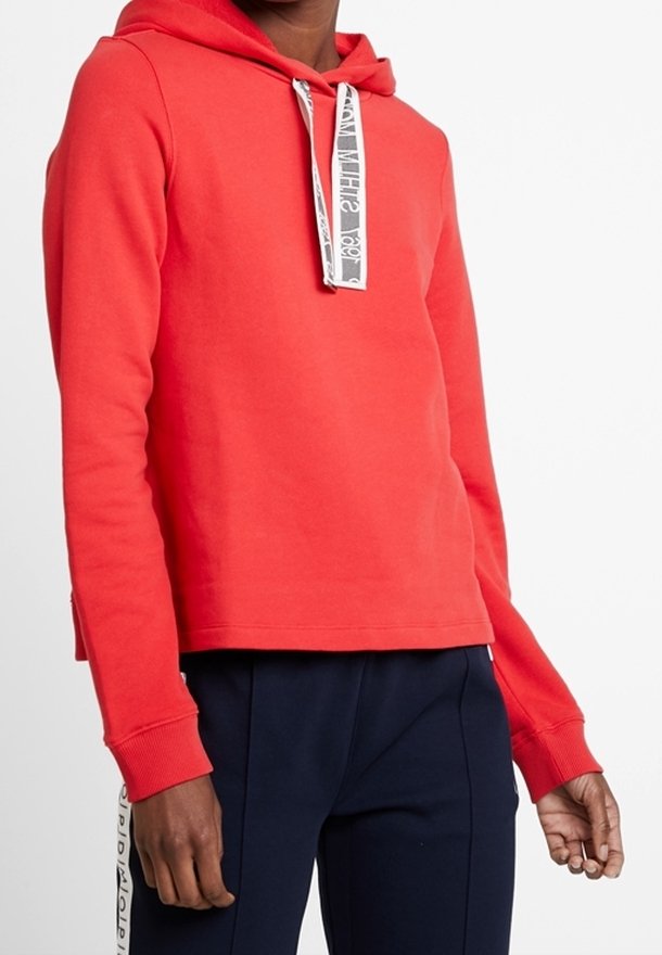 Personne portant un sweat à capuche rouge vif avec des cordons blancs et un pantalon bleu foncé avec une bande blanche sur le côté.