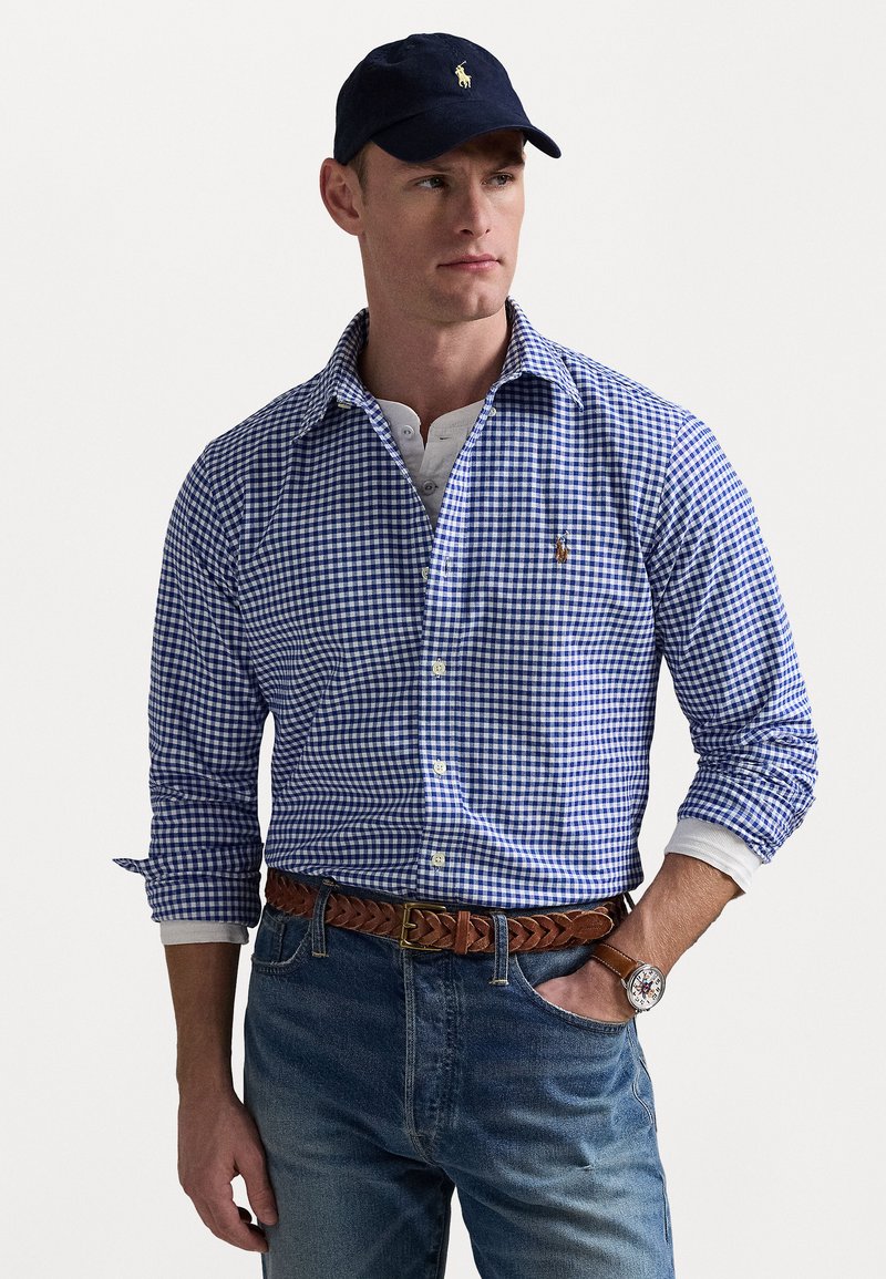 Blauw-wit geruit overhemd met een button-down kraag, gecombineerd met lichtblauwe jeans en een bruin gevlochten riem. Marineblauwe pet met logodetail.