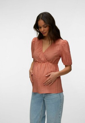 Femme enceinte portant un chemisier cache-cœur corail texturé et un jean bleu clair, tenant doucement son ventre, regardant vers le bas avec un doux sourire.