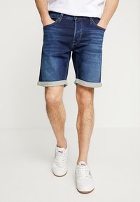 Indigo farkkushortsit käännetyillä lahkeilla, joissa on etutaskut ja suora malli. Yhdistetty valkoisiin lenkkitossuihin. Vähäinen varusteiden yksityiskohta.