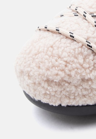 Chaussure en textile beige duveteuse avec une surface texturée, semelle extérieure noire et lacets à motifs. Forme arrondie, mettant en avant le confort et la chaleur.