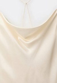Satin cami i mjuk krämfärg, med en draperad halsringning och delikata matchande band på baksidan. Slät, glänsande textur.