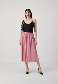 Svart sateng-camisole med tynne stropper, kombinert med en lys rosa plissert midi-skjørt. Brukt med svarte spisse hæler og en strukturert svart veske.
