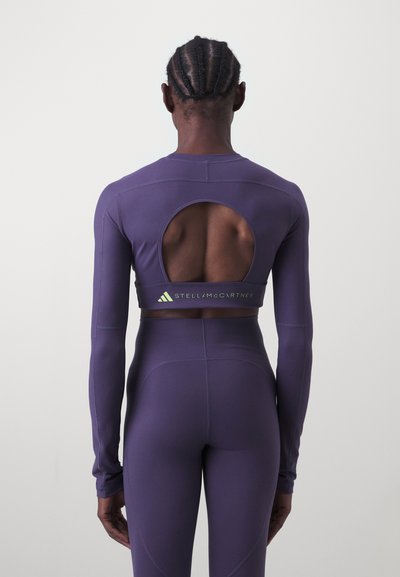 Fialová atletická mikina s dlhými rukávmi a veľkým výrezom na chrbte, s elastickým pásom s nápisom "Stella McCartney" v zelenej farbe. Hladká textúra.