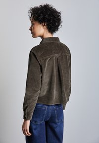 Chaqueta de pana marrón con cuello y puños abotonados, que presenta un yugo trasero y un corte relajado. Combinada con jeans de denim azul.