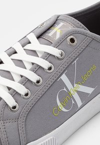 Grauer Segeltuch-Sneaker mit weißen Schnürsenkeln und Ösen. Auffällige Details sind ein großes "CK"-Logo und "Calvin Klein Jeans" in Gelb.