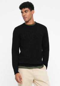 Maglione nero a maglia con logo "GUESS" in rilievo sul davanti, colletto rotondo e polsini a coste, indossato sopra una maglia verde scuro.