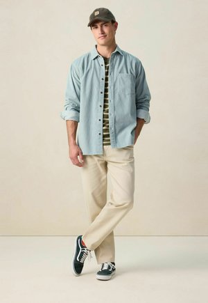 Chemise bleu clair à boutons par-dessus un t-shirt rayé vert, associée à un pantalon crème et des baskets vert foncé. La casquette arbore un logo.