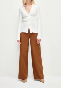 Gilet blanc à côtes avec un deep V-neck et un détail torsadé à l'avant, associé à un pantalon large marron et des chaussures à bout pointu beige.