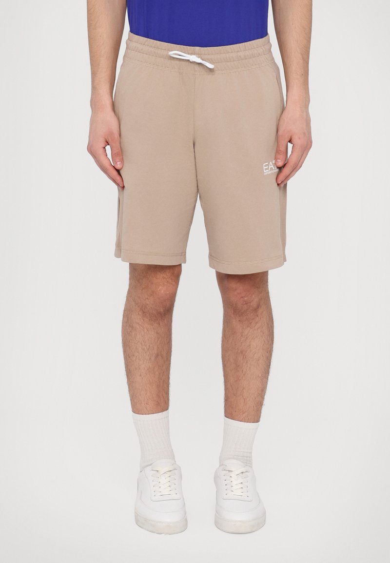 Beige bomullsshorts med elastisk liv og snøring. Sidelommer, kne-lengde, med en hvit logo på venstre hofte.