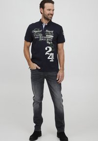 Marineblå polo med korte ermer med trykte logoer og tall; kombinert med mørkegrå jeans og svarte støvler.