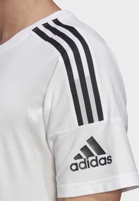 Bílé sportovní tričko s černým třípásovým designem na rameni a texturovaným logo Adidas na rukávu.