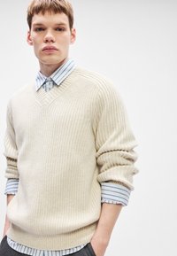 Ανοιχτό μπεζ πλέ knit πουλόβερ με V-λαιμό και ριμπ υφή, στρωμένο πάνω από ένα μπλε και λευκό ριγέ πουκάμισο με γιακά.