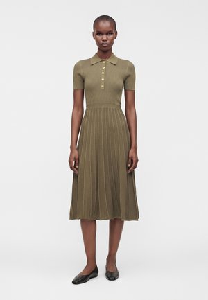 Donna con un vestito in maglia verde oliva a maniche corte con colletto, davanti abbottonato, gonna plissettata e scarpe nere basse, in piedi davanti a uno sfondo bianco.