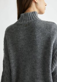 Maglione grigio a maglia con un alto colletto a coste, caratterizzato da motivi testurizzati e un piccolo logo sul retro. Materiale morbido e accogliente.
