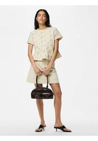 Femme portant un haut crème à manches courtes en broderie anglaise et une jupe assortie, tenant un petit sac à main marron foncé, portant des sandales noires à talons, debout.
