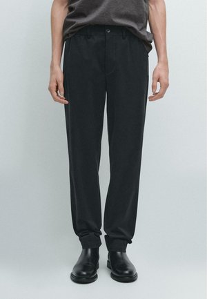 Pantaloni neri su misura in tessuto liscio con cintura, tasche frontali e orli risvoltati, abbinati a scarpe nere alla caviglia.