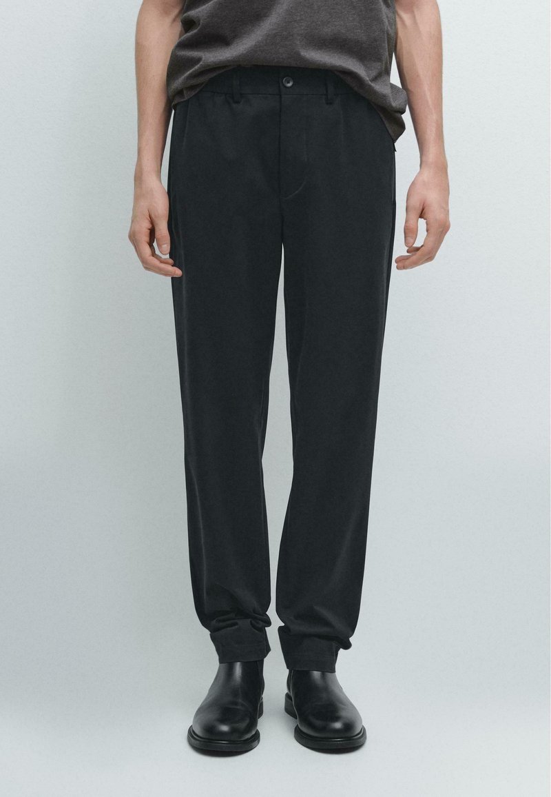 Schwarze, maßgeschneiderte Hose aus glattem Stoff mit Bund, Vorderentaschen und umgeschlagenen Säumen, kombiniert mit schwarzen Schuhen bis zum Knöchel.