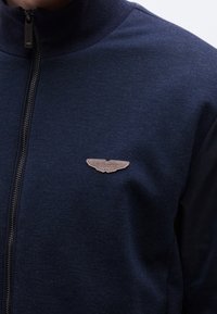 Gros plan d’une personne portant une veste zippée bleu marine avec un petit logo beige en forme d’aile Aston Martin sur la poitrine.