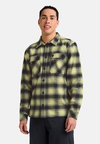 Chemise en flanelle à carreaux jaunes et noirs, avec un devant boutonné, deux poches poitrine et une coupe décontractée. Tissu épais avec un motif texturé.