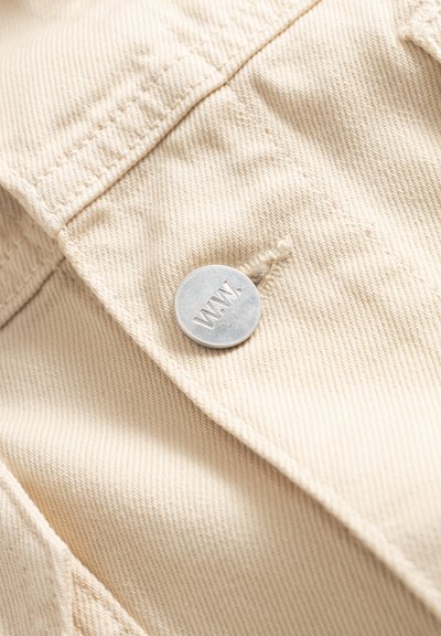 Beige fabric with visible stitching and a round silver button engraved with the letters "W.W." attached near a buttonhole.