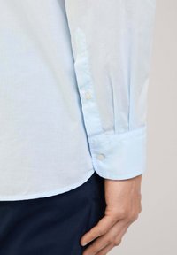 Camisa de manga larga azul claro, fabricada en un material suave, con puños abotonados y un dobladillo ligeramente redondeado. Presenta sutiles frunces en los hombros.