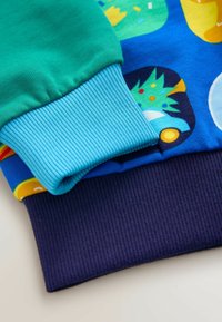 Farverige sweatermanchetter har tekstureret ribbet stof i grøn, lys blå og marineblå, med livlige trykte mønstre på kroppen.