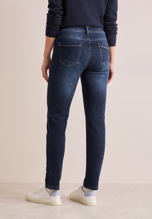 Dunkelblaue Jeans im Slim Fit, mit genähten Gesäßtaschen und einem mittelhohen Bund. Getragen mit weißen Sneakers.