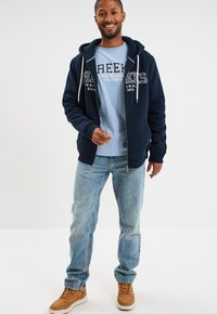 Sudadera con cremallera color marino, con un forro suave, cordones grises, combinada con una camiseta de color azul claro y jeans de denim azul deslavado. Zapatillas marrones completan el look.