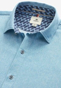 Camisa de rayas azul claro con botones, con un forro de cuello con patrón en contraste azul marino y dos botones en la tapeta frontal. Etiqueta de la marca visible.