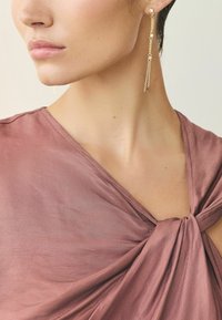 Gold chain earrings with dangling accents, paired with a mauve fabric top featuring a knotted detail and smooth texture.