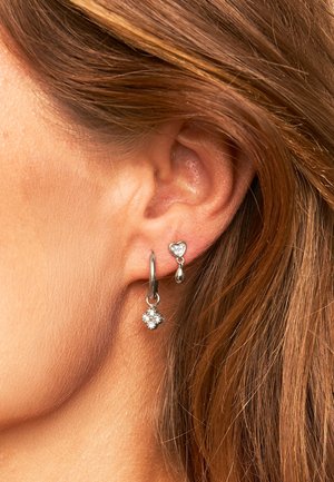 Silberne Ohrringe mit einem runden Ring, einem kubischen Kristallanhänger und einem kleinen herzförmigen Charme, im Kontrast zu braunem Haar.