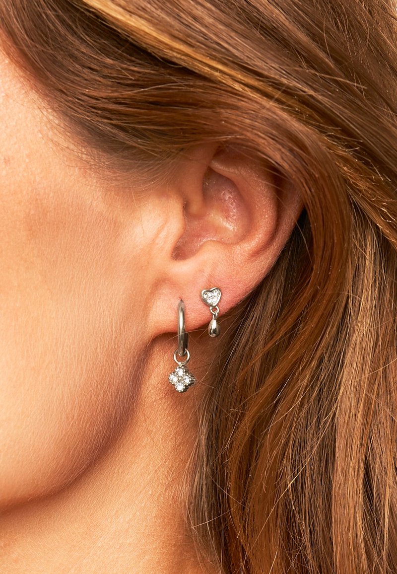 Srebrne kolczyki z okrągłą obrączką i podwieszoną kryształową zawieszką w kształcie serca, umieszczone na tle brązowych włosów.