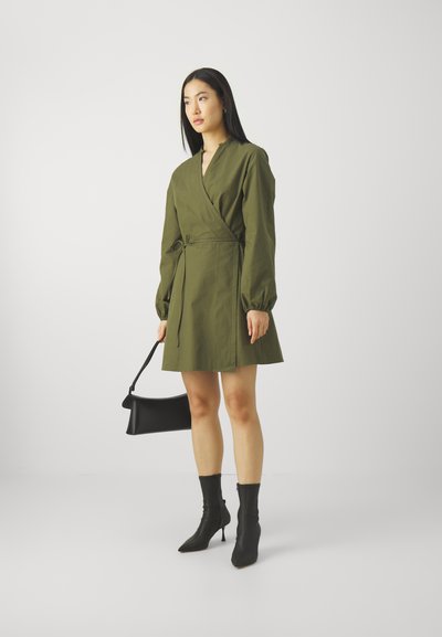 Robe portefeuille olive avec des manches longues, une taille ceinturée et un décolleté en V. Portée avec des bottines noires et un sac à main noir.
