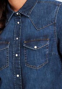 Denim shirt in dark blue with a pointed collar, front snap buttons, two chest pockets, and contrast stitching. Soft texture and structured fit.