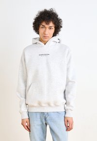 Sweat à capuche gris en tissu doux, avec une poche kangourou, des poignets côtelés et le texte "STUDIO EDITION" imprimé sur la poitrine.