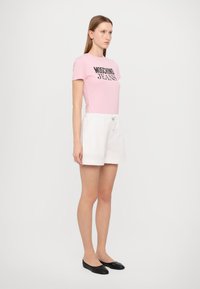 Ružová bavlnená tričko s čiernym nápisom "Moschino Jeans", spárované s bielymi šortkami a čiernymi balerínkami. Model stojí na jednoliatom pozadí.