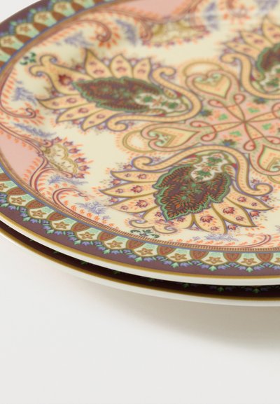 Pile d'assiettes en céramique ornées de motifs floraux et géométriques complexes dans des tons roses pâles, verts et marron sur un fond blanc.