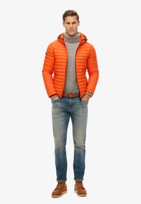 Orange quiltet pufferjakke med hætte, grå turtleneck, lyseblå jeans og brune snørebåndsstøvler. Jakken har sort lynlås og detaljer.