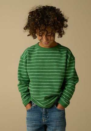 Kind mit lockigem Haar, trägt grünen gestreiften Pullover und blaue Jeans, Hände in den Taschen, schaut nach unten, neutraler Hintergrund.